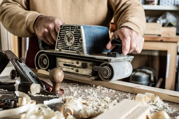 Electric belt sander, sanding machine in male hand. Processing of workpiece on light brown wooden table. Side view, close up