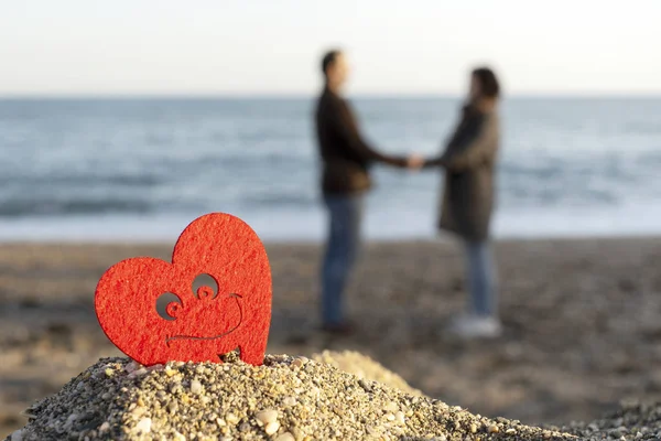 Red heart on a mountain of sand by the sea with a couple of lovers in the background. concept of san valentine — Stock Photo, Image