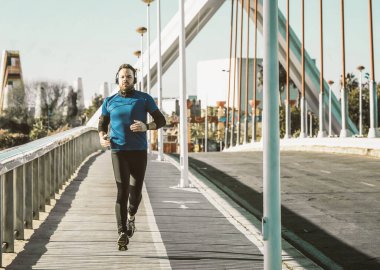 Genç adam mavi gömlekli bir köprüde koşuyor ve kulaklıkla müzik dinliyor..