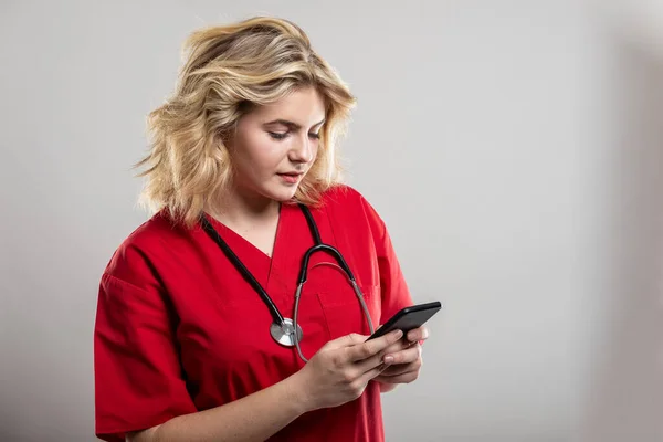 Portrait of nurse wearing red scrub texting on smartphone on studio gray background with copy space advertising area