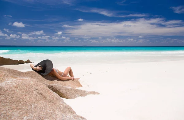 Praia de areia tropical com água azul-turquesa, bela mulher deita na rocha em chapéu de sol preto . — Fotografia de Stock