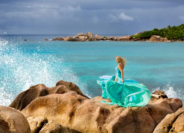 在塞舌尔 美丽的金发碧眼的女人在一些奇异的岩石之上 在汹涌的印度洋海浪旁边 玩着她美丽的蓝绿色华丽的长裙 图库照片