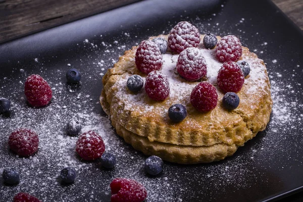 Kuchen mit Himbeeren, Blaubeeren, Sanddorn bestreut Puderzucker auf einem schwarzen Teller — Stockfoto