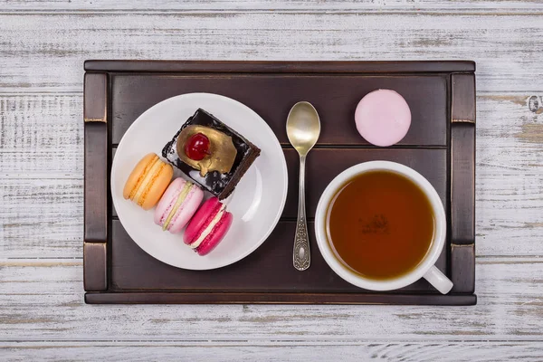 Cup of tea, piece chocolate cake and macaron cakes on tray on white wooden table. Lifestyle concept. — Stock Photo, Image