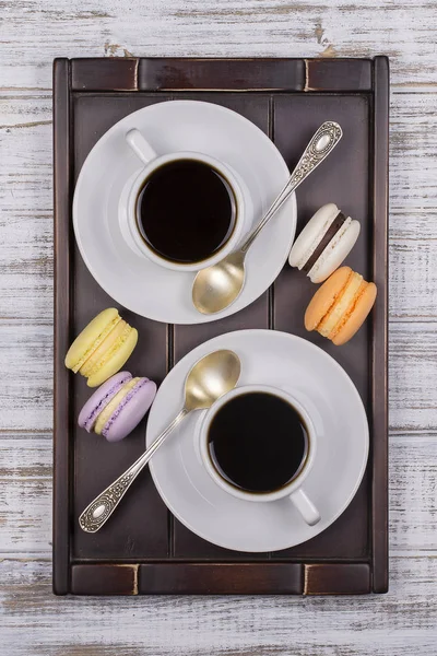 Duas xícaras de café, colher e bolos de macaron na bandeja na mesa de madeira branca. Conceito de estilo de vida. Vista superior — Fotografia de Stock