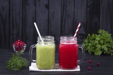 İki cam kupa suyu güler yüzlü sarsıntısını yeşil maydanoz, dereotu, brokoli, avokado ve kırmızı siyah ahşap arka plan üzerine kızılcık