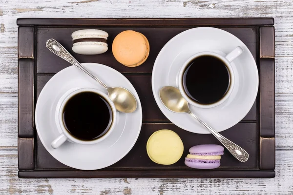 Duas xícaras de café, colher e bolos de macaron na bandeja na mesa de madeira branca. Conceito de estilo de vida. Vista superior — Fotografia de Stock