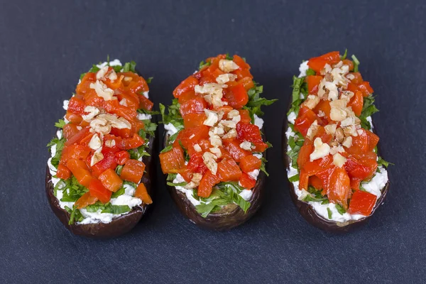 Tres berenjenas rellenas de queso crema, hierbas verdes, pimientos rojos y amarillos a la parrilla sobre un fondo de pizarra negra, de cerca — Foto de Stock