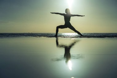 Silhouette of woman standing at yoga pose on the tropical beach during sunset. Girl practicing yoga near sea water