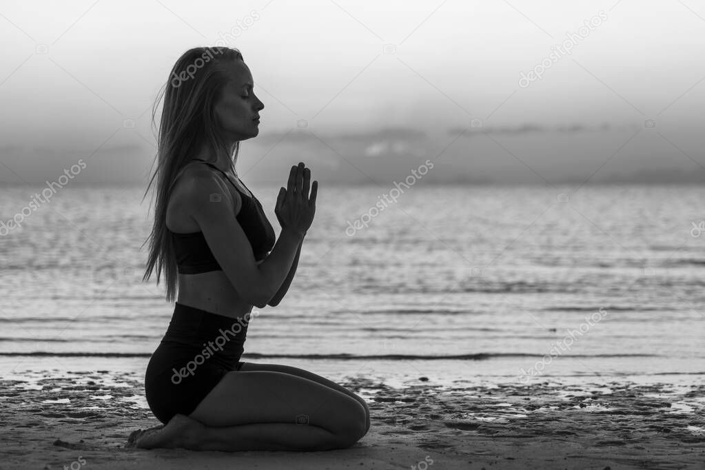 Silhouette of woman sitting at yoga pose on the tropical beach during sunset. Caucasian girl practicing yoga near sea water. Black and white