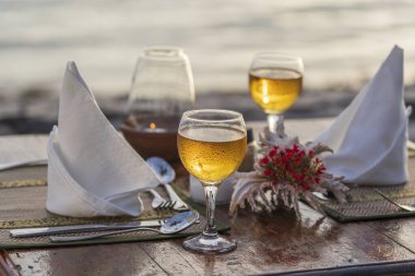 Gün batımında tropikal sahilde, deniz kenarındaki ahşap masada iki şarap kadehi. Romantik akşam yemeği konsepti