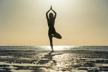 Gün batımında tropik plajda yoga pozu veren kadın silueti. Beyaz kız deniz suyunun yanında yoga yapıyor.