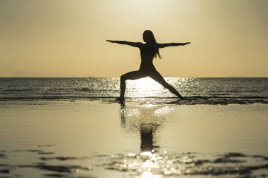 Gün batımında tropik plajda yoga pozu veren kadın silueti. Beyaz kız deniz suyunun yanında yoga yapıyor.