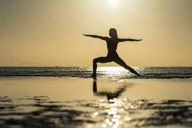 Gün batımında tropik plajda yoga pozu veren kadın silueti. Beyaz kız deniz suyunun yanında yoga yapıyor.