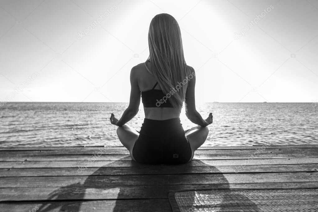 Silhouette of woman sitting at yoga pose on the tropical beach during sunset. Caucasian girl practicing yoga near sea water.
