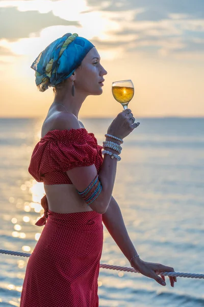 热带海滩上 一个可爱的女人在海浪边 面对美丽的落日 举着一杯白葡萄酒 女孩正在喝着一杯葡萄酒欣赏日落 休闲和旅行的概念 — 图库照片