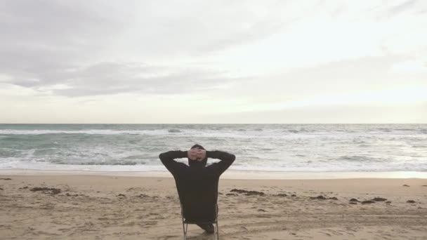 Um homem senta-se numa cadeira no meio da praia — Vídeo de Stock