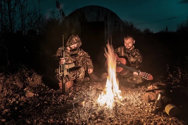 two military men with guns and guitar sitting by fire in post apocalyptic world