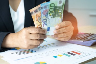 Avro banknotları tutan, çalışan, hesaplayan ve analiz eden Asyalı muhasebeci rapor projesini hesap makinesi ve grafik raporuyla modern ofiste: finans ve iş konsepti.