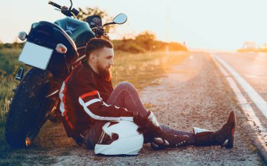 Motosikletine uzanmış güzel sakallı yakışıklı beyaz şoför. Sol elini gün batımına karşı beyaz kaskın üzerine koyuyor.