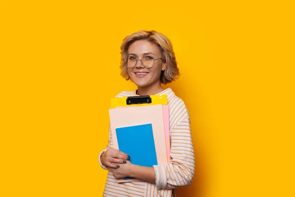 Preciosa mujer rubia caucásica felizmente sonriendo sobre un fondo amarillo mientras sostiene algunos libros — Foto de Stock