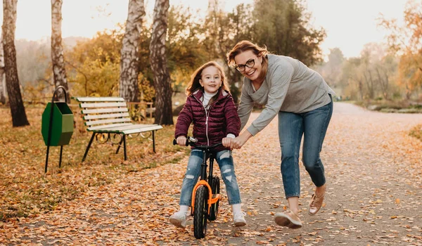 Güneşli bir sonbahar akşamında küçük ve gülümseyen kızına bisiklet sürmeyi öğreten neşeli bir anne. — Stok fotoğraf