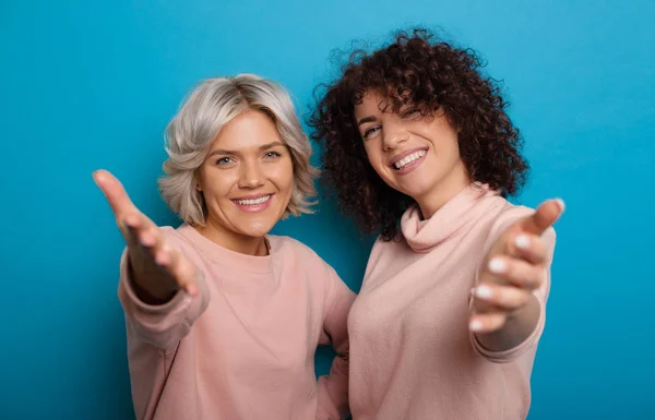 Dos sonrientes damas caucásicas con el pelo rizado señalándote como una invitación sobre fondo azul — Foto de Stock