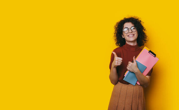 Lovely curly haired student holding some books is recommending something by posing on a yellow blank spaced background and showing the approbation sign