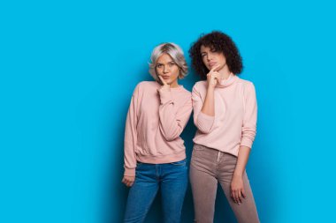 Two caucasian sisters dressed in pink sweaters and jeans are looking thoughtful posing on a blue background clipart