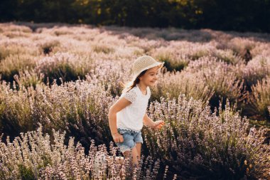 Güzel bir şapka takan gülümseyen beyaz kız güneşli bir yaz gününde lavanta tarlasında neşeyle oynuyor.
