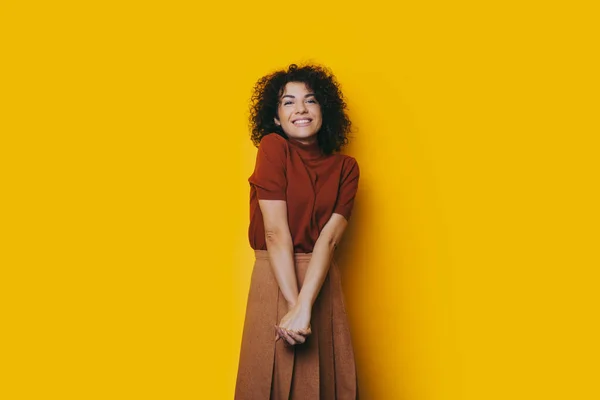 Bella ragazza caucasica con i capelli ricci e sorriso allegro in posa in un bel vestito con le mani incrociate su uno sfondo giallo — Foto Stock