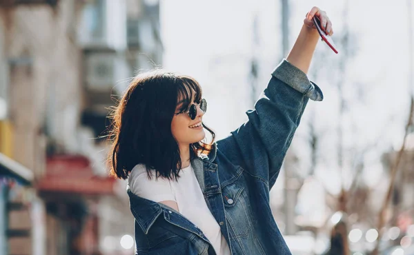 Atemberaubendes kaukasisches Mädchen mit schwarzem Haar, das lächelt und durch ihre Brille schaut, während es mitten in der Stadt ein Selfie macht — Stockfoto