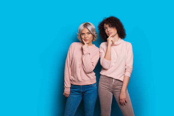 Two caucasian sisters dressed in pink sweaters and jeans are looking thoughtful posing on a blue background