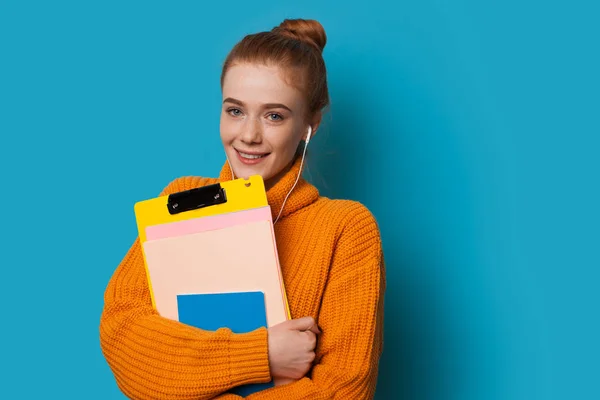 Χαριτωμένο χαμογελαστός μαθητής με κόκκινα μαλλιά και φακίδες κρατά κάποια βιβλία που είναι ντυμένοι σε ένα πορτοκαλί πουλόβερ, ενώ θέτουν σε ένα μπλε φόντο και ακούγοντας — Φωτογραφία Αρχείου