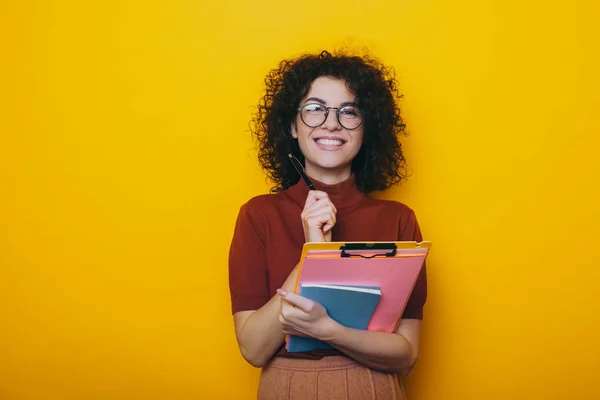 Curly young caucasian girl wearing eyeglasses is holding a pen and some books posing on a yellow background — Stock Photo, Image