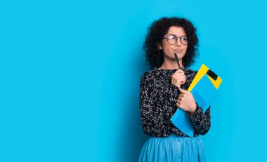 Kıvırcık saçlı, beyaz, çekici bir esmer elinde bir kalem ve birkaç kitap tutarken mavi, boş bir arkaplanda bir şeyler düşünüyor.