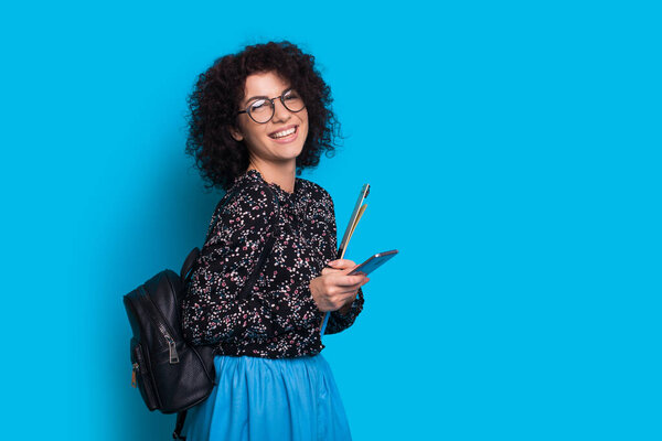 Lovely caucasian student with curly hair and eyeglasses holding a mobile and some sheets is posing on a blue background