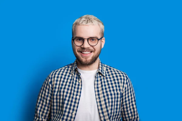 Menino bonito com cabelo loiro e barba bonita está olhando através de seus óculos enquanto posando com confiança no fundo azul — Fotografia de Stock