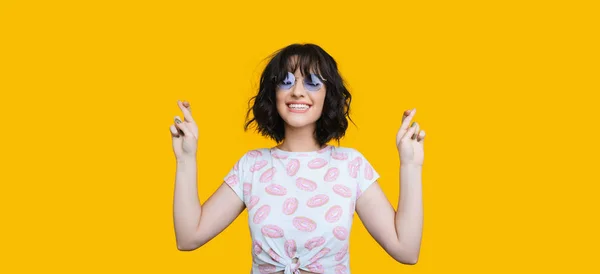 Vrolijk brunette Kaukasisch meisje draagt bril poseert met gesloten ogen en gekruiste vingers op een gele achtergrond — Stockfoto