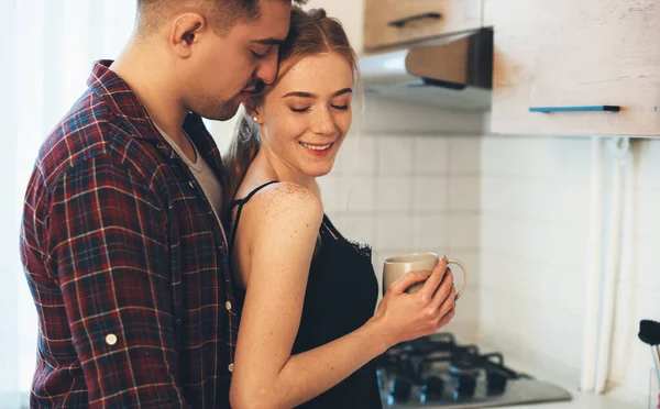 Uomo attento abbracciare la sua ragazza caucasica con lentiggini e capelli rossi mentre beve un caffè in cucina — Foto Stock