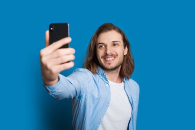 Kahverengi sakallı uzun saçlı beyaz adam mavi arka planda telefonunu kullanarak kendi portresini yapıyor.