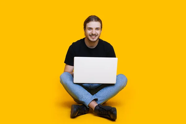 Caucasiano homem sem barba com cabelos longos sentado com pernas cruzadas digitando algo no computador enquanto posando em um fundo amarelo — Fotografia de Stock