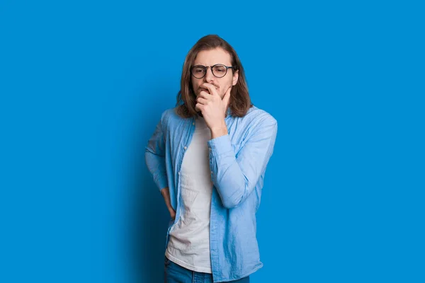 Homem caucasiano com cabelos longos e barba agradável está tocando seu queixo enquanto olha para o lado através de seus óculos e posando no fundo azul — Fotografia de Stock