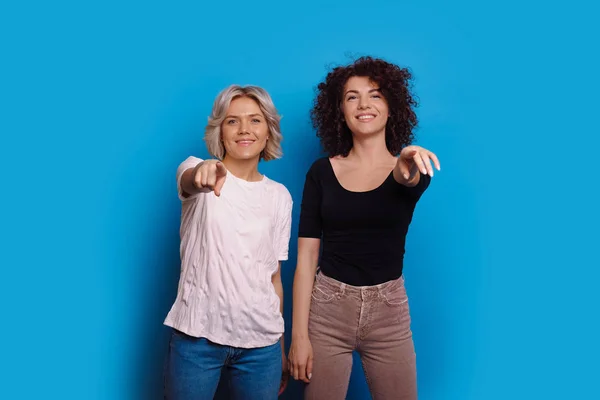 Duas mulheres caucasianas alegres vestidas de jeans com cabelo encaracolado estão apontando para a câmera enquanto sorrindo em um fundo azul — Fotografia de Stock