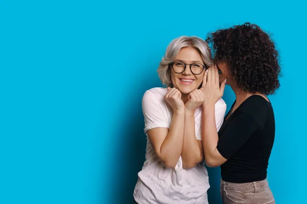 Lockiges kaukasisches Mädchen spricht etwas Geheimnisvolles zu ihrer blonden Freundin, die eine Brille trägt und auf einem blauen Leerzeichen-Hintergrund posiert — Stockfoto