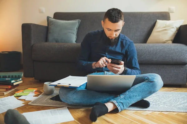 Ocupado freelancer caucásico con una camisa y barba bonita está trabajando duro sentado en el suelo con un ordenador portátil y algunos documentos —  Fotos de Stock