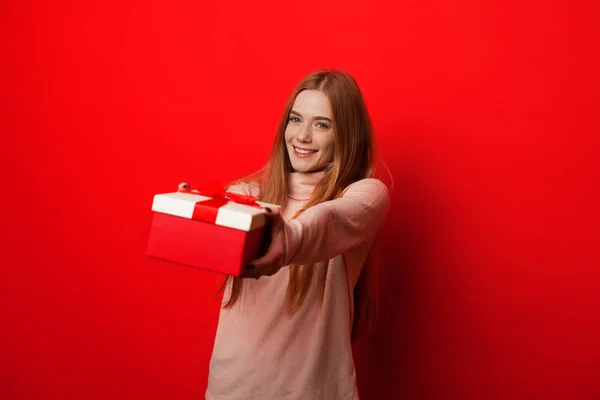 Alegre chica caucásica con el pelo rojo y pecas está sosteniendo un regalo mientras posa sobre un fondo rojo — Foto de Stock