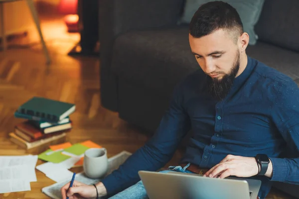 Snygg kaukasisk entreprenör med stort skägg arbetar på sin bärbara dator medan du skriver något och dricka en kaffe — Stockfoto