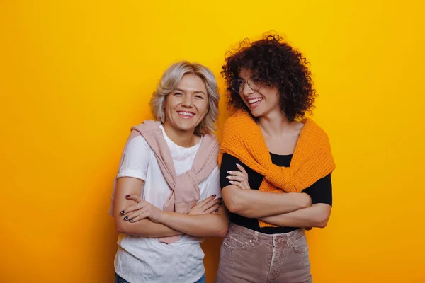 Zwei hübsche kaukasische Frauen mit lockigem Haar posieren selbstbewusst mit verschränkten Händen auf gelbem Hintergrund, lächeln und haben Spaß — Stockfoto