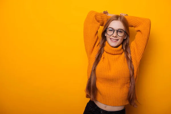 Encantadora estudiante pelirroja con el pelo rojo y gafas está estirando sus manos mientras posa en un suéter naranja sobre un fondo amarillo — Foto de Stock